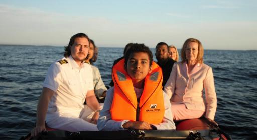 ilmstill der Videoprojektion mit Rettungsboot, den Hauptfiguren und einem Jungen mit Schwimmweste auf dem Meer. Im Hintergrund der Horizont und die aufgehende Sonne.